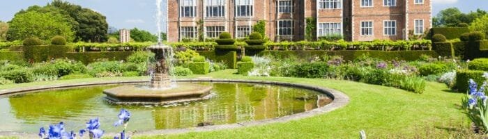 Elegant historic mansion with manicured gardens and fountain under a bright blue sky.