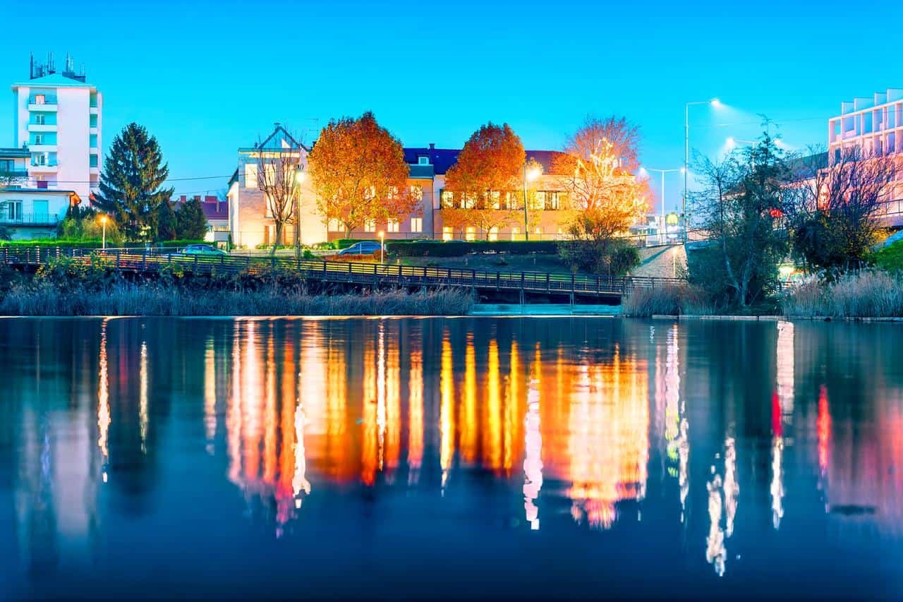 Colorful cityscape at dusk reflected in river, vibrant lights and trees, urban scenery for travel and vacation photography.
