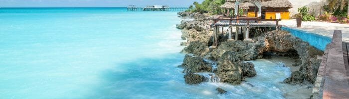 Turquoise ocean water crashing against rocky shoreline with thatched-roof cabanas and palm trees at tropical beach resort.