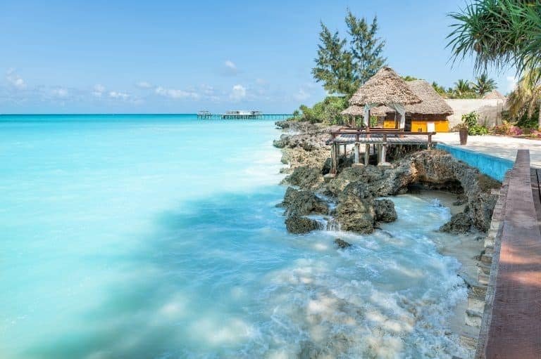 Turquoise ocean water crashing against rocky shoreline with thatched-roof cabanas and palm trees at tropical beach resort.