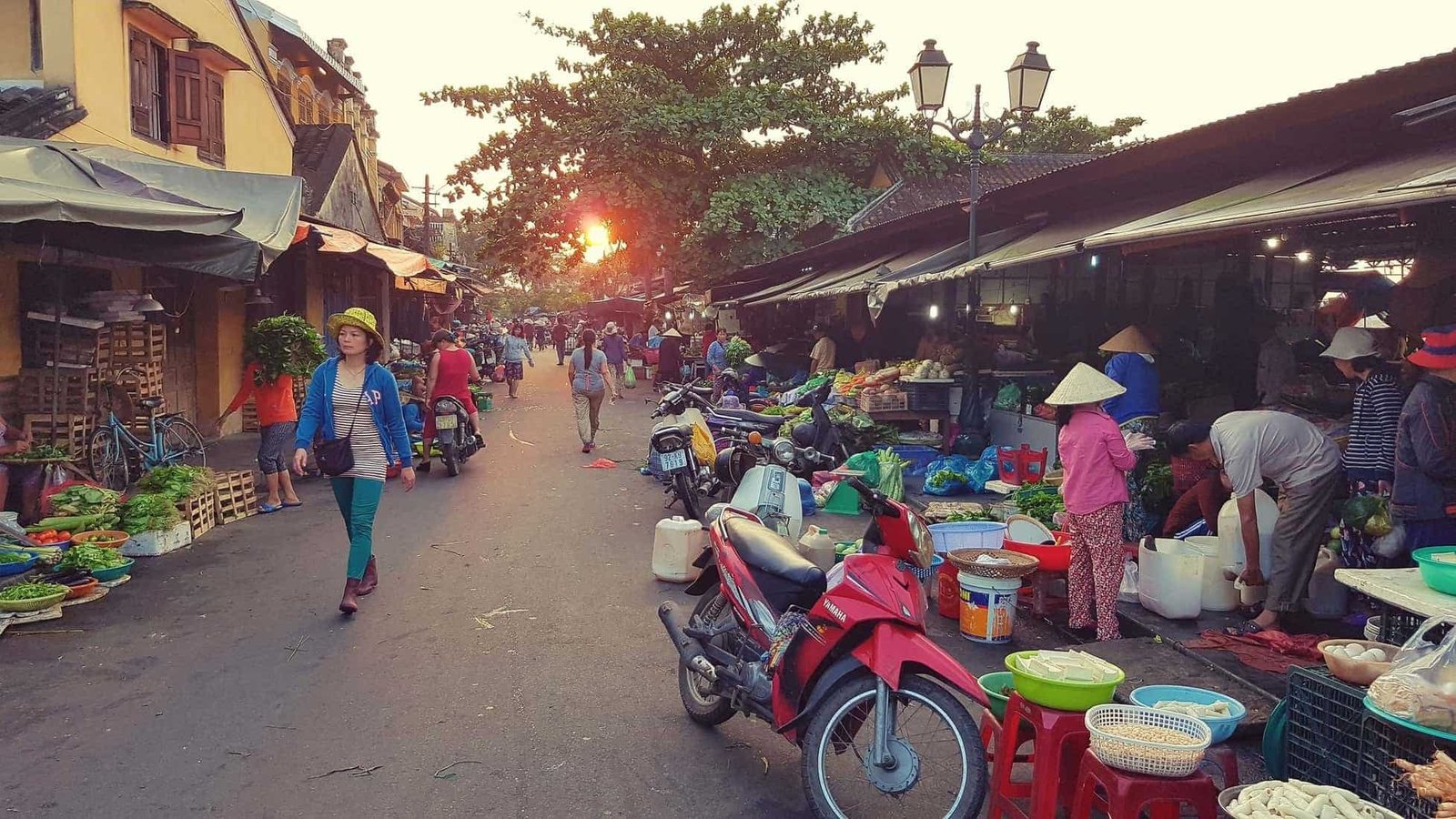 Street market in Hoi An Vietnam with foods in plastic bowls, goods for sale piled high and motorbikes parked outside the stalls each covered by a protective overhead awning (blind).