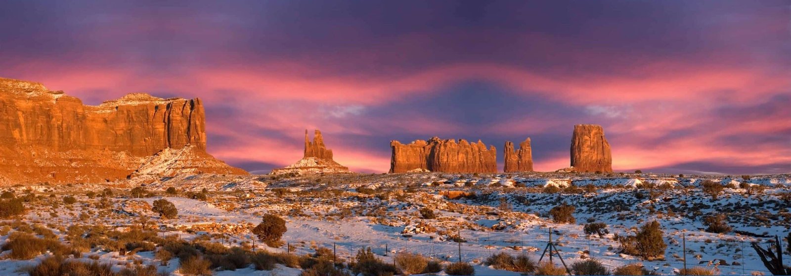 Panoramic sunset scene from the Monument Valley Navajo Indian Reservation Tribal Park in Arizona and Utah. Monoliths rise above the desert and glow as the setting sun shines upon them. Winter snow adds a surreal effect. The area is famous for the old John Ford movies being filmed here, many of them starring John Wayne. Monument Valley is now an international travel destination with people and tourists coming from all over the world just to look at the view.