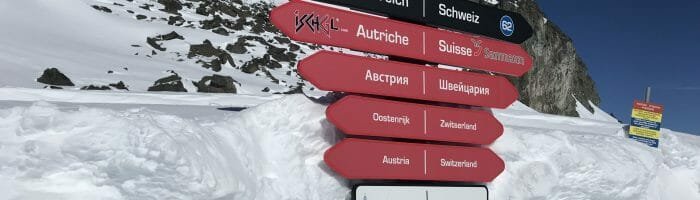 Snowy mountain border sign in the Alps, indicating Austria, Switzerland, and other countries, under a clear blue sky.