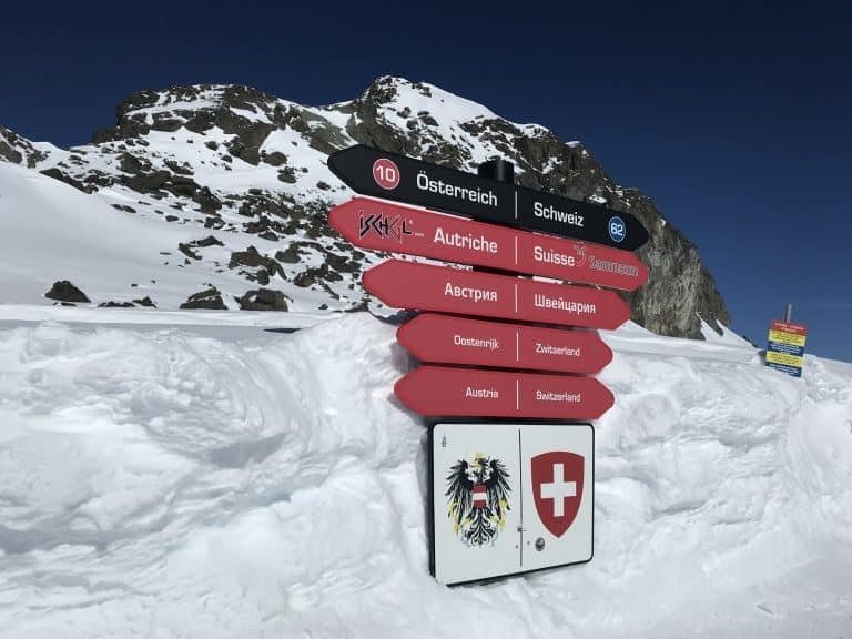 Snowy mountain border sign in the Alps, indicating Austria, Switzerland, and other countries, under a clear blue sky.