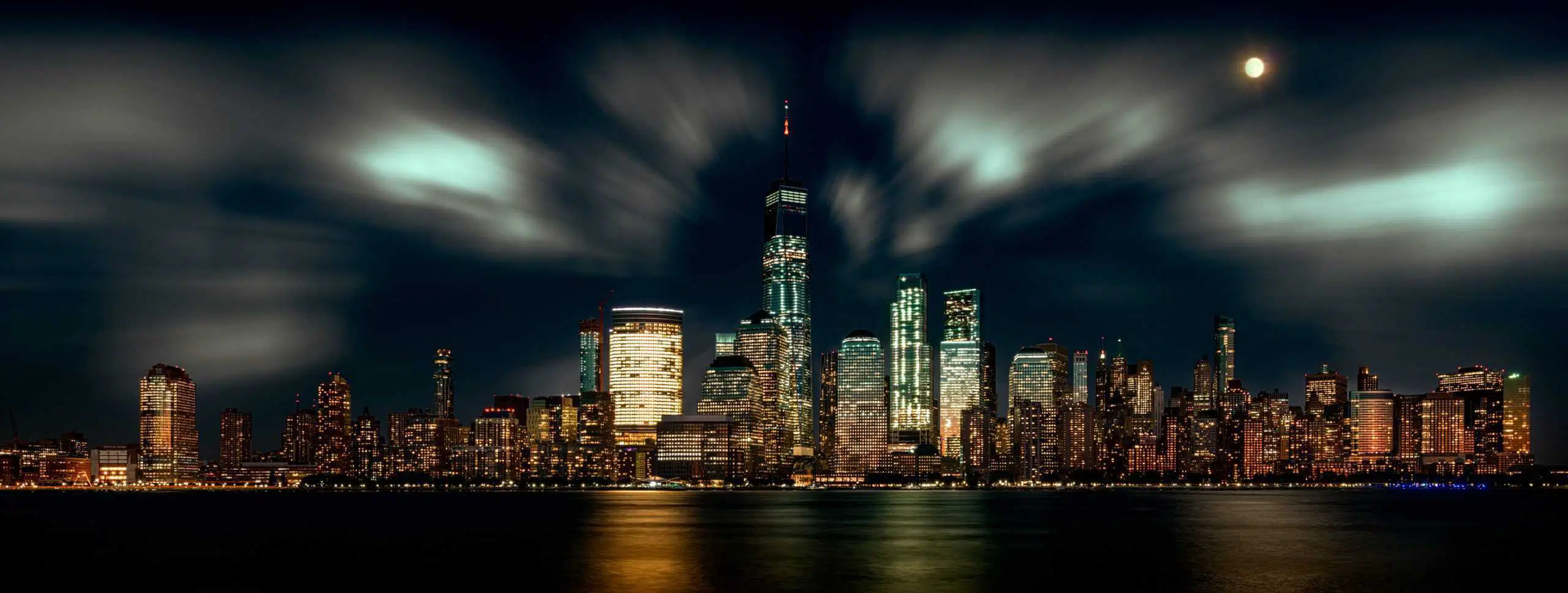 Vibrant New York City skyline illuminated at night with moving clouds and city lights reflecting on water.