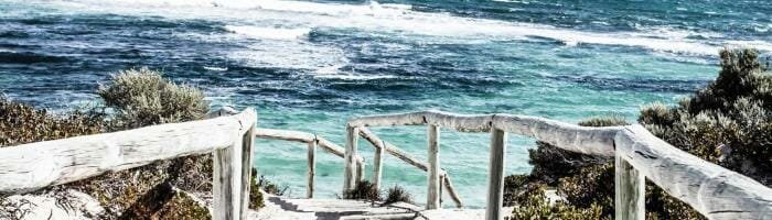 Bright sandy beach with white stairs and weathered wooden railing overlooking turquoise ocean waves.