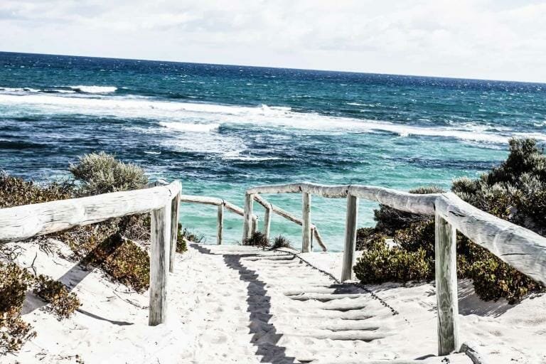 Bright sandy beach with white stairs and weathered wooden railing overlooking turquoise ocean waves.