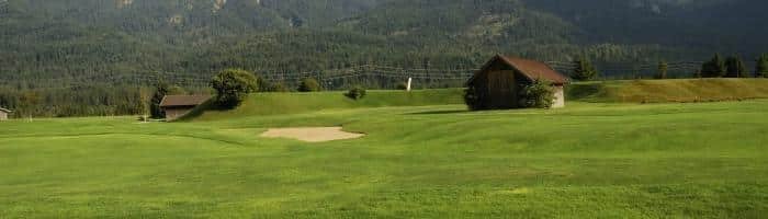 Mountain golf course in scenic landscape with lush green fairways and wooden cabins, surrounded by forested mountains.