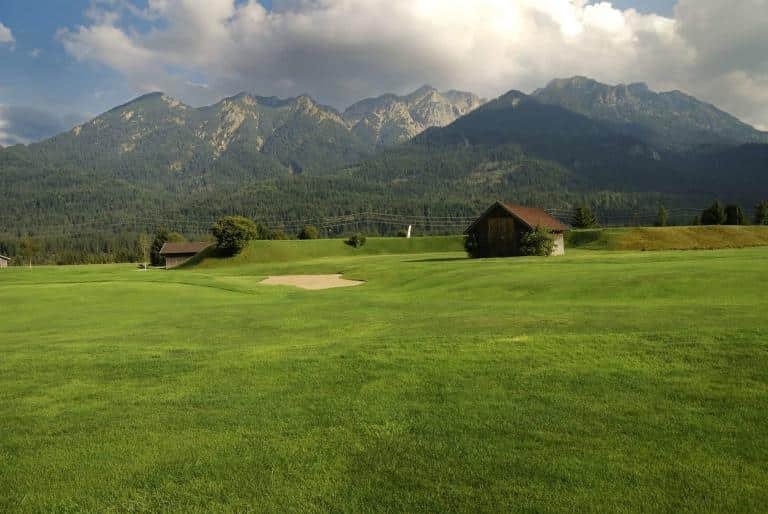 Mountain golf course in scenic landscape with lush green fairways and wooden cabins, surrounded by forested mountains.