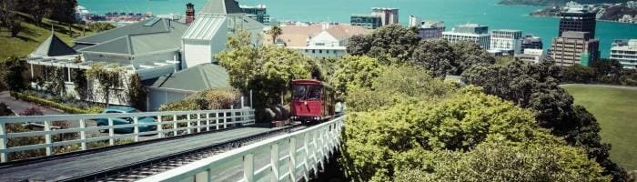 Vintage red funicular train ascending through lush greenery in Wellington, New Zealand, overlooking the city skyline and harbor.