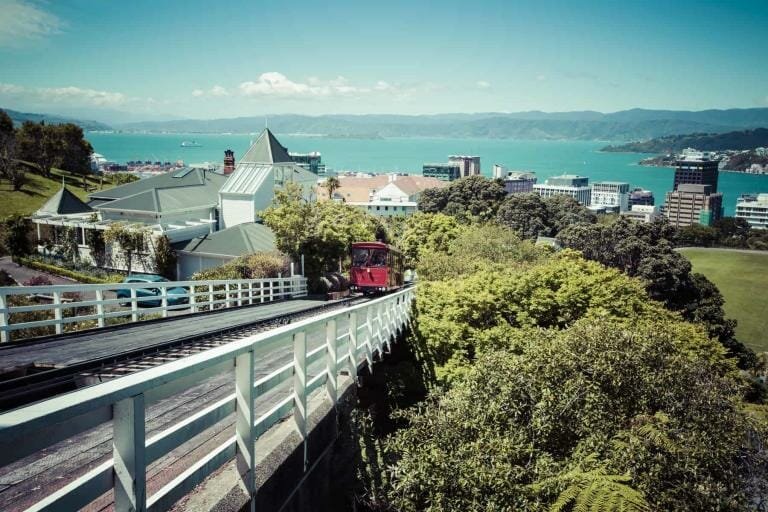 Vintage red funicular train ascending through lush greenery in Wellington, New Zealand, overlooking the city skyline and harbor.