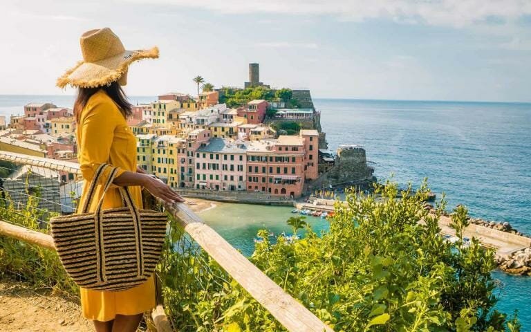 Colorful coastal village in Italy with a woman in a yellow dress and sun hat enjoying scenic ocean views.