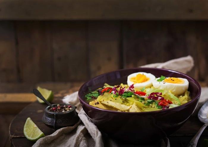 Freshly prepared chicken noodle soup with boiled eggs, fresh vegetables, and lime wedges on a rustic wooden table.
