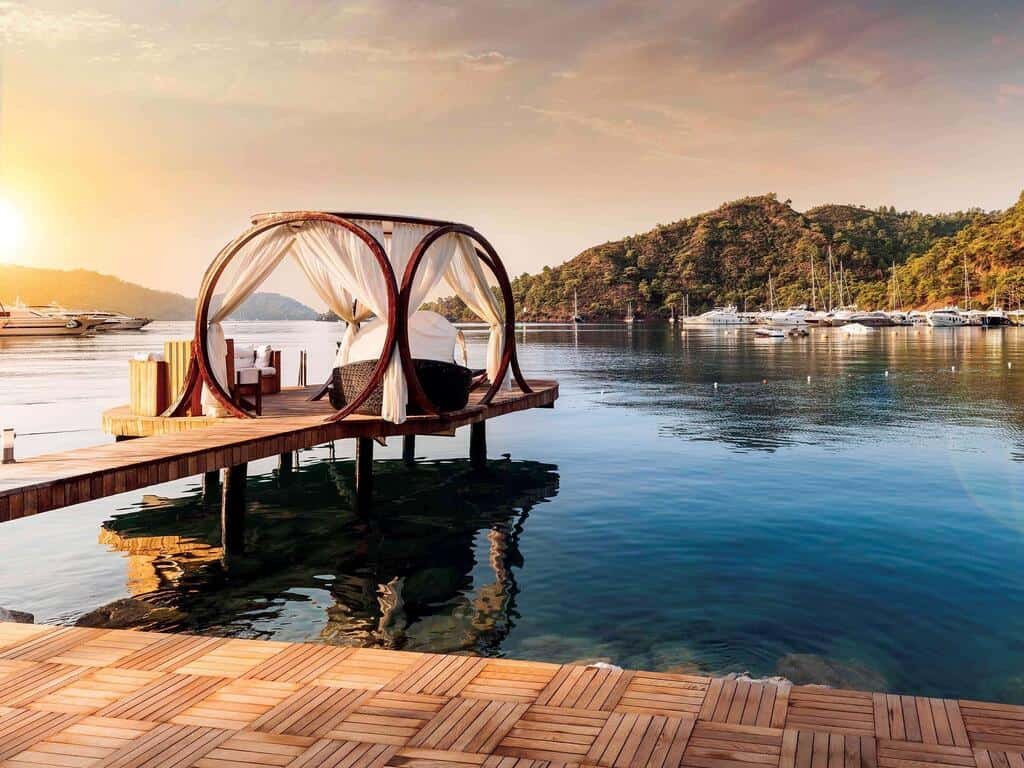 Secluded water bungalow with canopy bed on a wooden pier at sunset, overlooking calm marina and lush green hills.