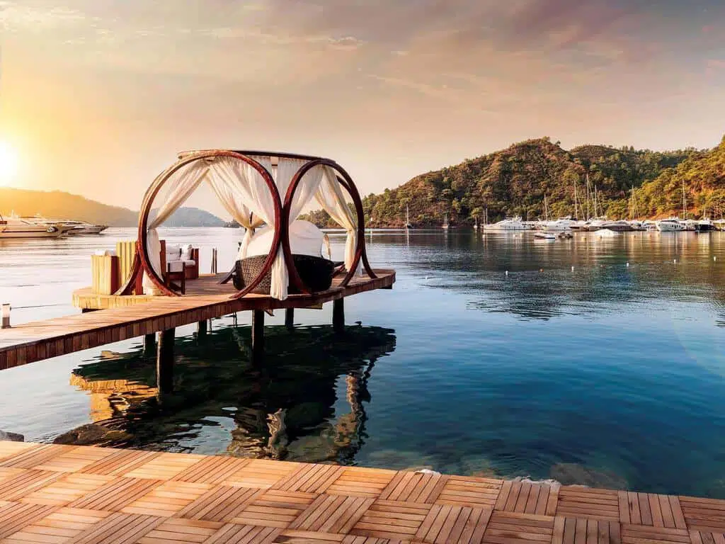 seaside hotels turkey near — Secluded water bungalow with canopy bed on a wooden pier at sunset, overlooking calm marina and lush green hills.