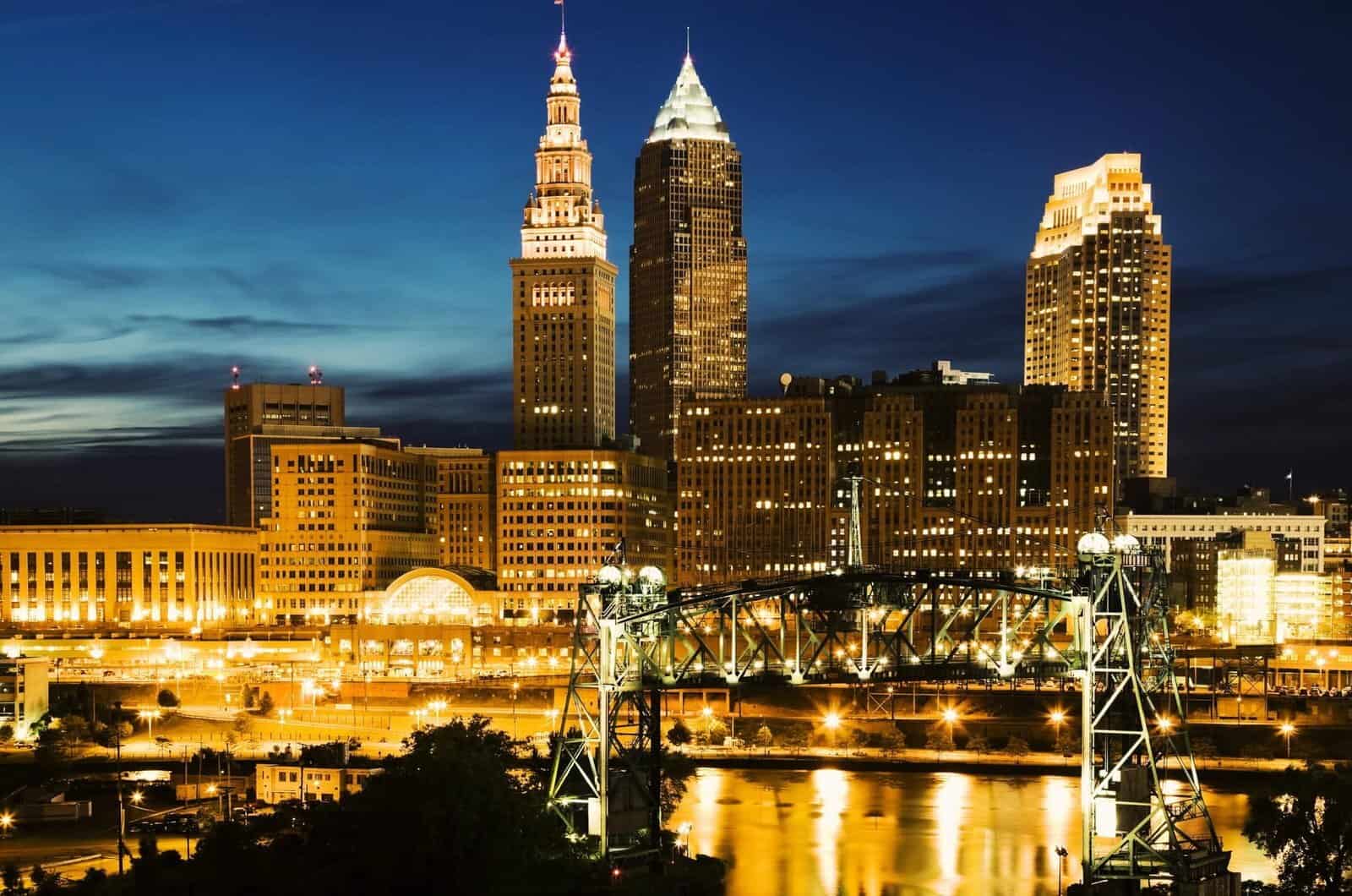 Skyline of Cleveland at night with illuminated buildings and historic steel bridge reflecting on the river.