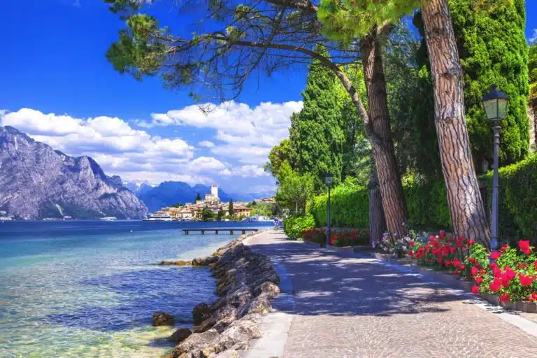 Serene lakeside promenade with colorful flowers and tall trees against a backdrop of mountains and a charming lakeside town.