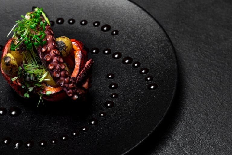 Fresh grilled octopus appetizer with microgreens on black textured plate, fine dining seafood dish.
